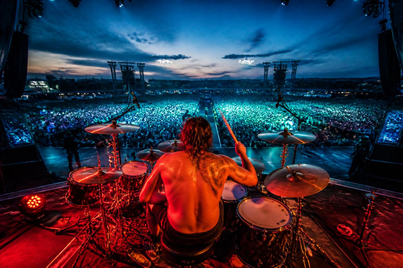 A stadium rock show seen from behind the band, looking out at a packed crowd and blazing stage lights